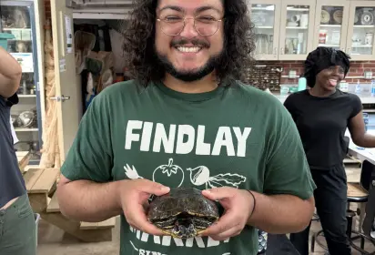 Smiling man in a green shirt holding a box turtle. Woman in the background laughing.