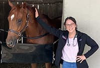 Claire wearing a stethoscope outside a stall with a brown horse at Belterra Racetrack Clinic.
